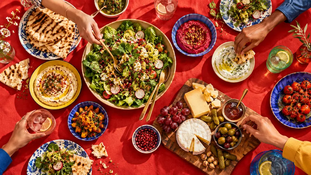 Overhead view of a generous sharing spread of colourful salads, dips and flatbreads on a tomato-red tablecloth