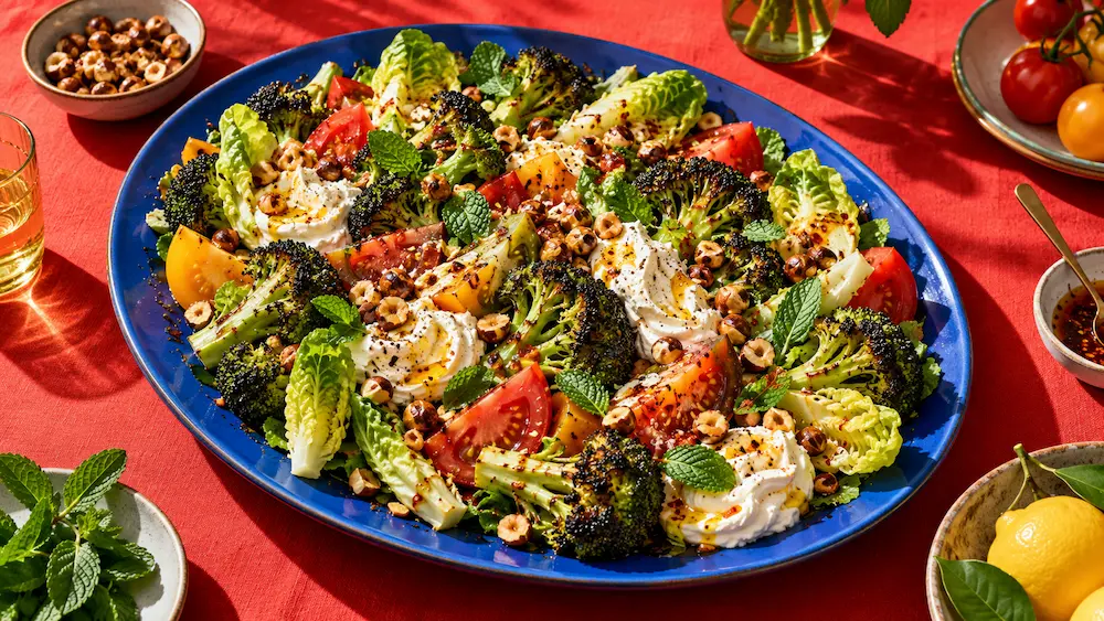 A huge, colourful salad of charred broccoli, torn leaves, tomatoes and whipped feta on a tomato-red table