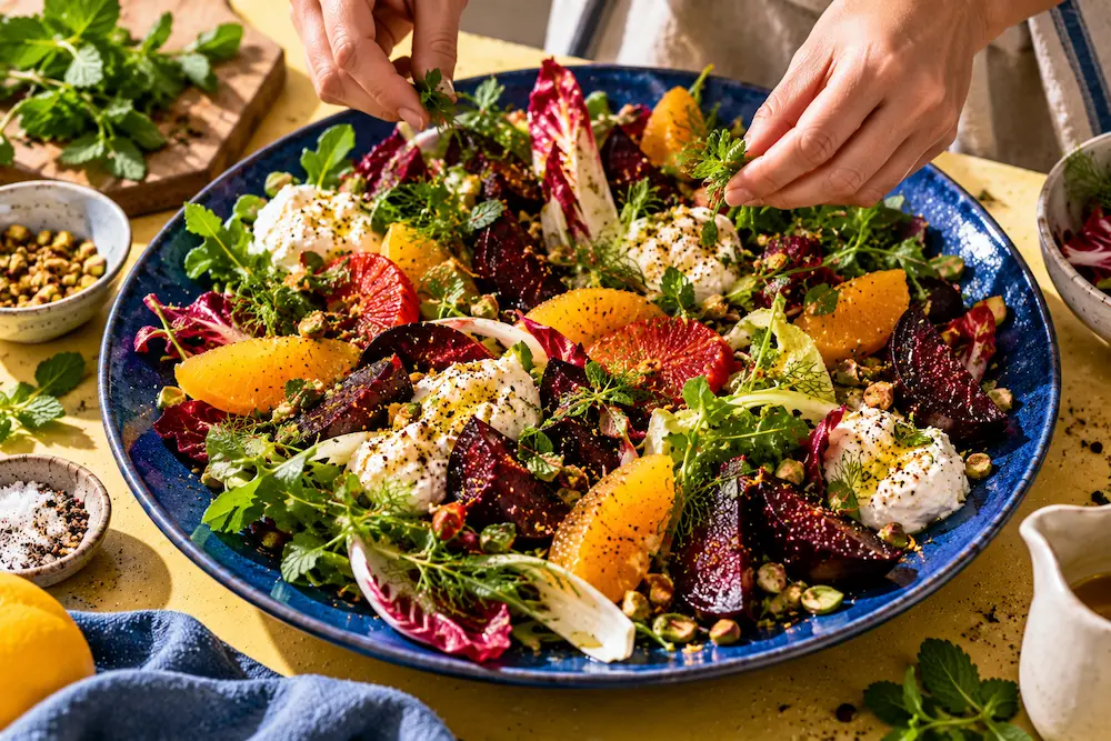 Hands placing the last torn herbs on a large salad arranged on a wide cobalt platter with space between the ingredients