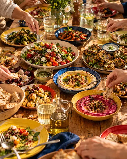 A long tasting table with plates of finished dishes and hands reaching in to share