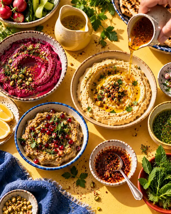 Bowls of hummus, beetroot labneh and smoky aubergine dip swirled with olive oil on a butter-coloured table